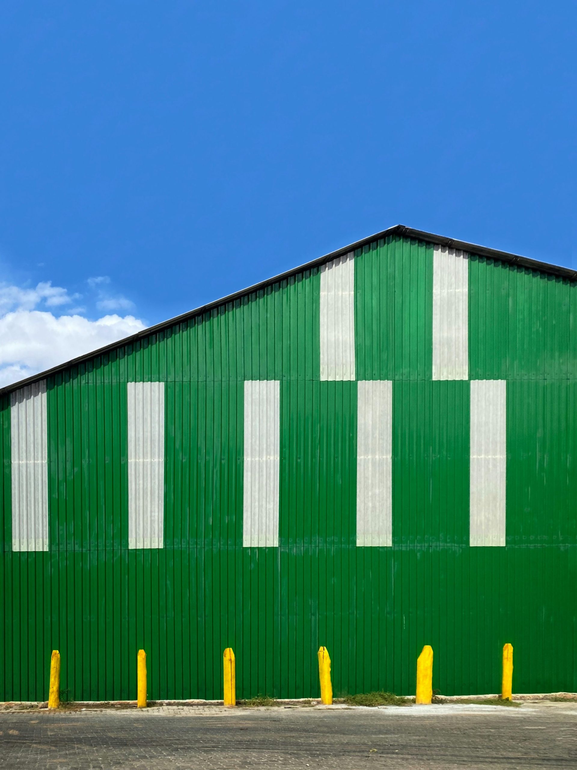 Toma vertical de un almacén de color verde intenso con rayas blancas y postes amarillos, bajo un cielo azul despejado.